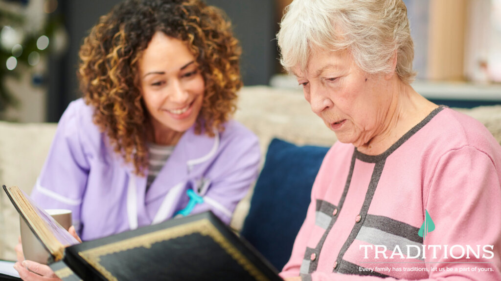 A close-up of a caregiver and elderly woman looking at a photo book on the couch. The caregiver is wearing a purple top and the elderly woman is wearing a pink sweater. Traditions Logo on the bottom right corner.